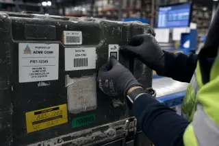 Technician's gloved hands attaching an RFID tag to a shipping crate amid a busy warehouse with labels and conveyor in the background.
