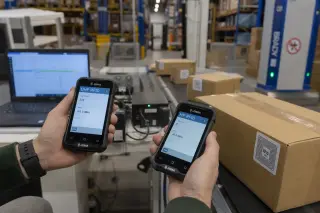 Engineer’s hands comparing two RFID readers over a conveyor belt in a warehouse, with RFID-tagged boxes, shelves, and laptop in the background.