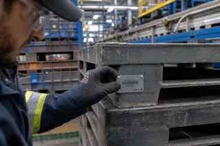 Technician's gloved hand attaching a metal RFID tag to a steel pallet in a warehouse with industrial equipment in the background.