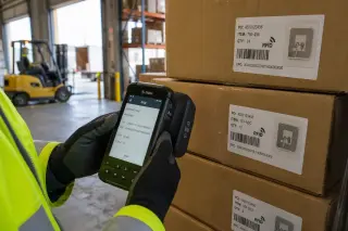 Technician's hand scanning a carton with RFID reader on conveyor belt in warehouse, both RFID and barcode visible, industrial setting with cool lighting.